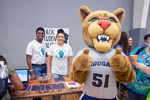 Scooter the Cougar holding thumbs up in the foreground while students gather around in the background.