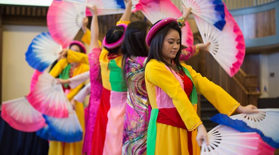 Women dancing traditional dance, wearing brighly colored dresses and waving pink and blue fans.