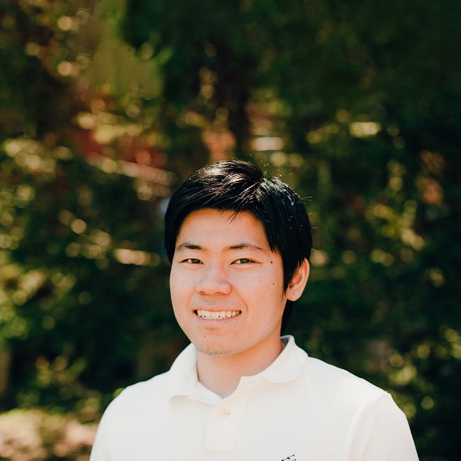 International: Male student standing in front of greenery, smiling at the camera.