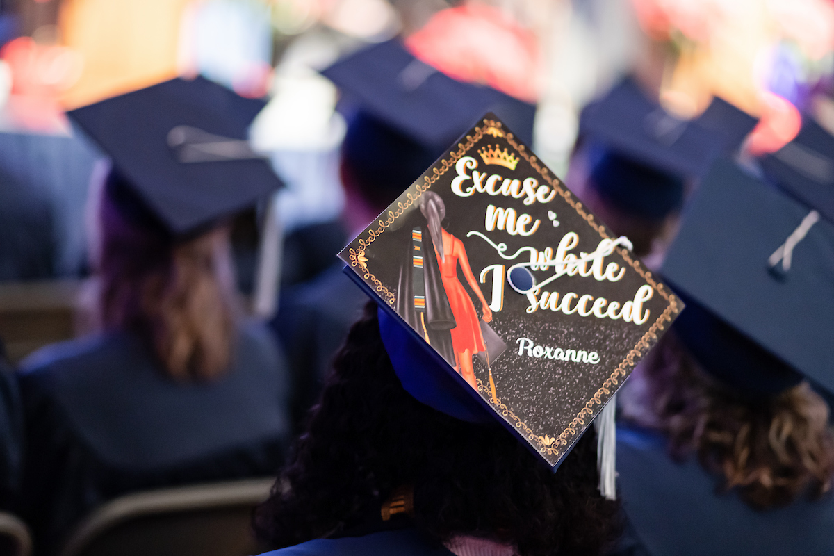 Commencement ceremony with graduate cap reading "Excuse me while I succeed - Roxanne".