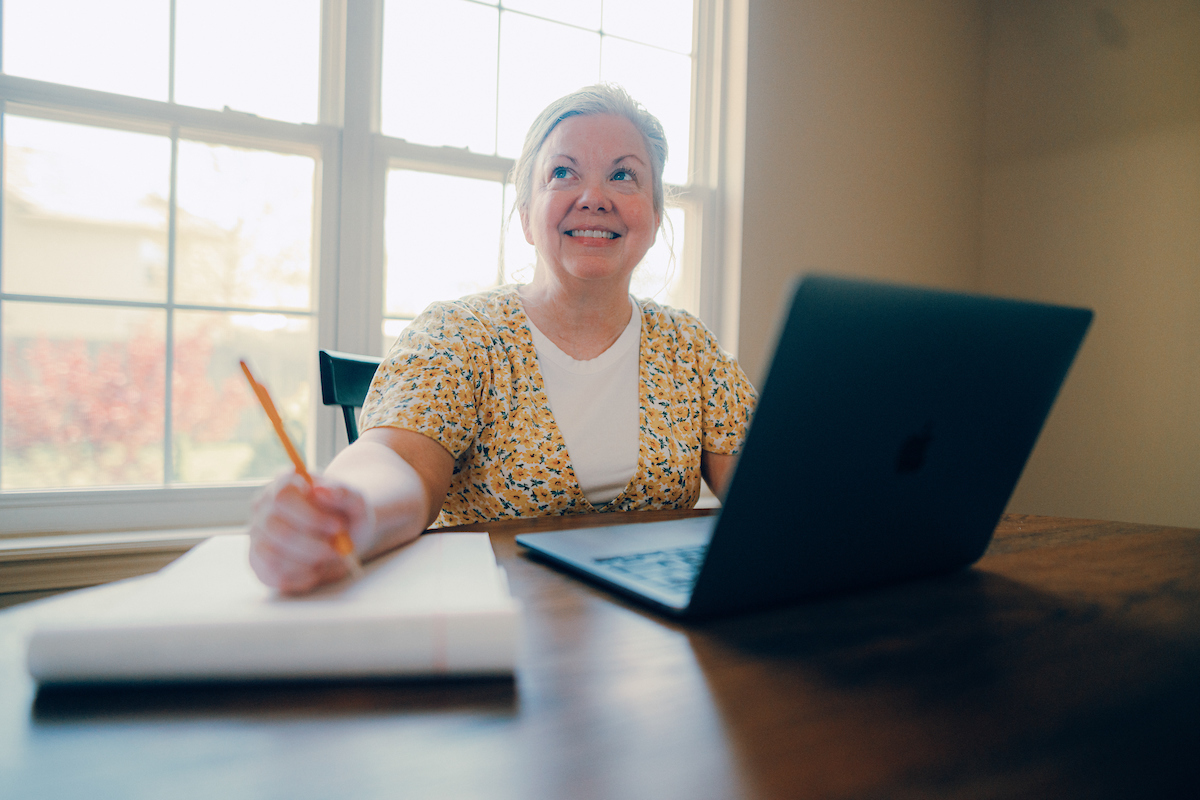 Adult student sitting at dining table in front of window, with a laptop and writing pad in front of her.