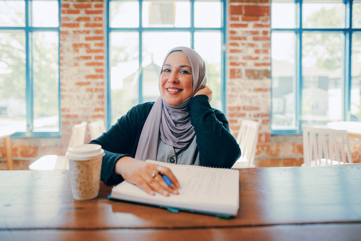 Woman wearing a head scarf sitting at a table with a notepad and writing utensil.