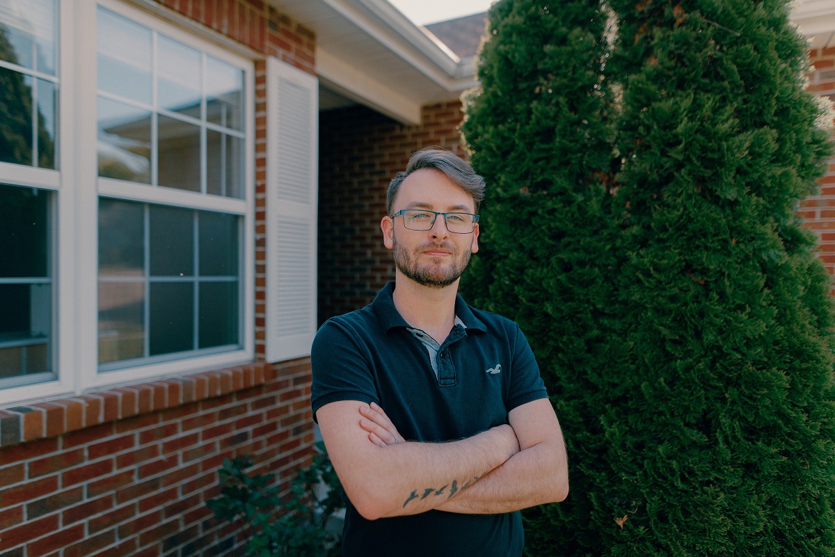 Robert W. Plaster School of Business: Man wearing glasses with arms crossed standing in front of a brick building and shrubs.