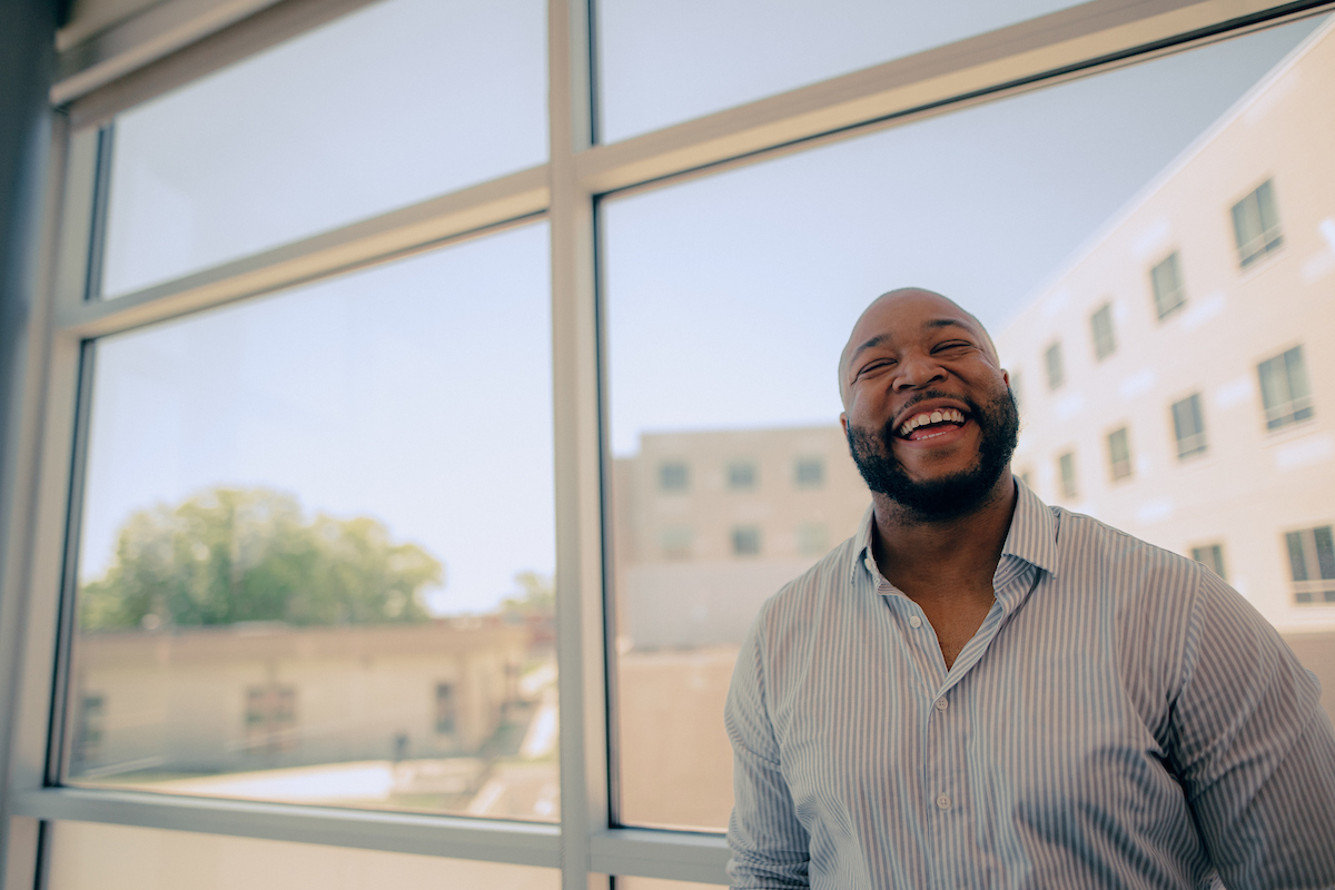 Graduate: Man laughing while facing the camera and standing in front of a window.