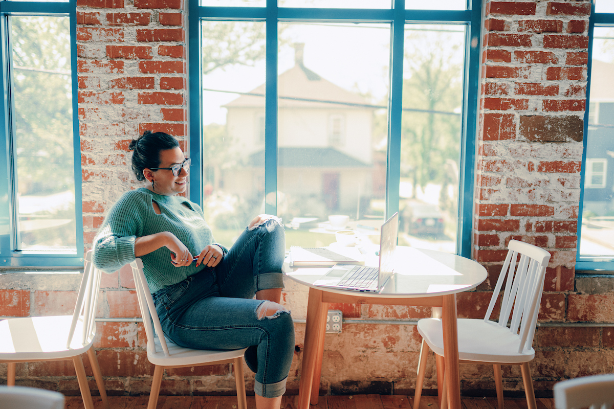 Adult student sitting at table in front of window, viewing a laptop while sitting with one leg pulled up on the chair.