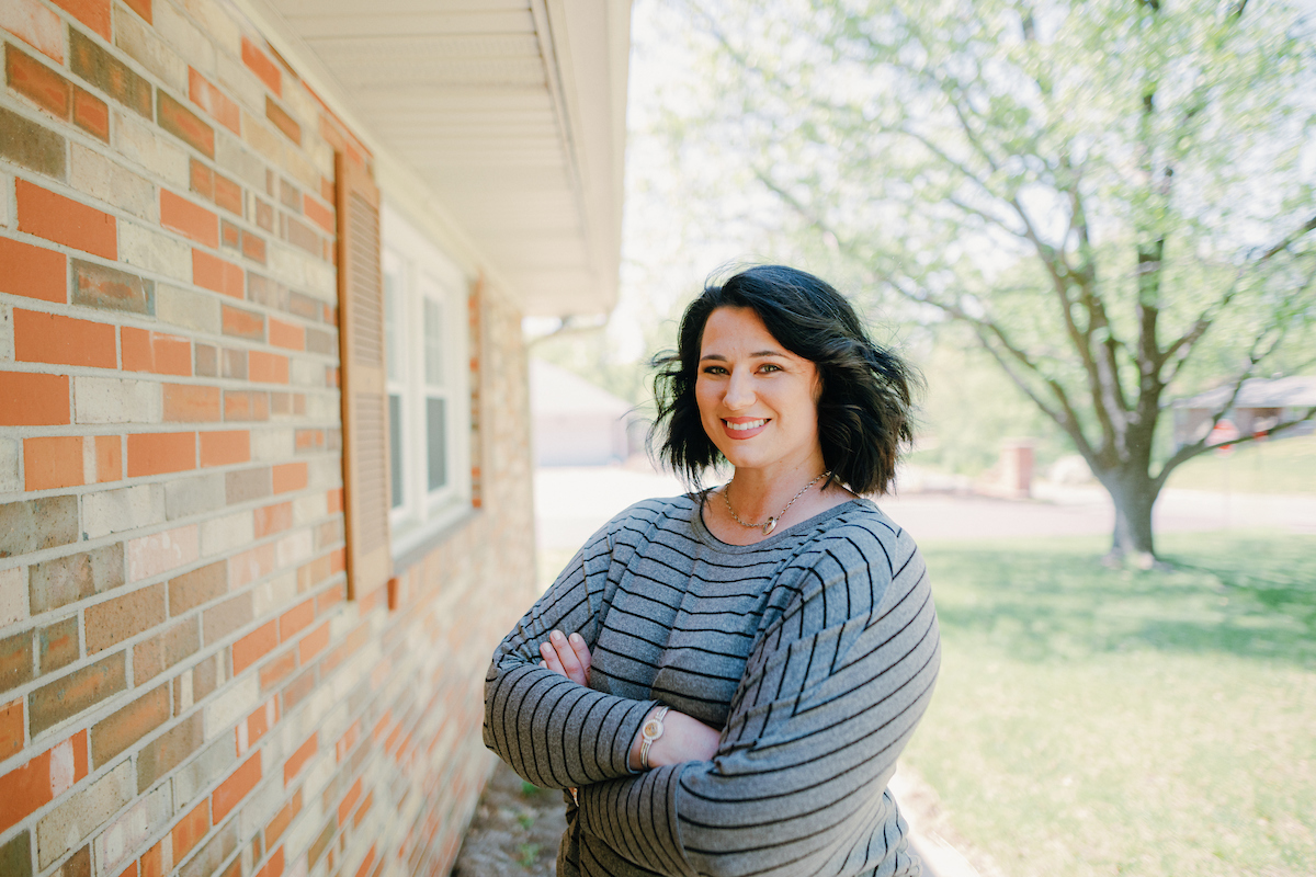 For Nursing Programs: Woman standing outside of a brick house, with arms crossed and looking at the camera.