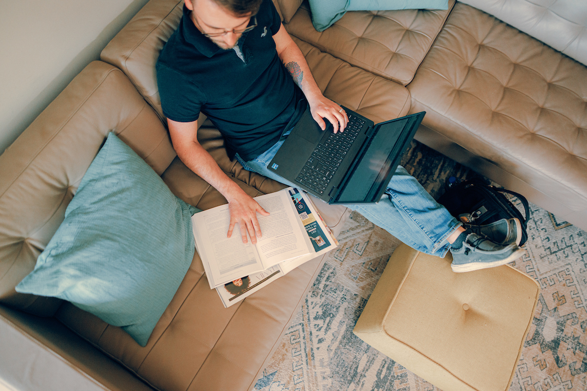 Man sitting on a couch with a laptop on his lap and a book open next to him, with his feet propped up.