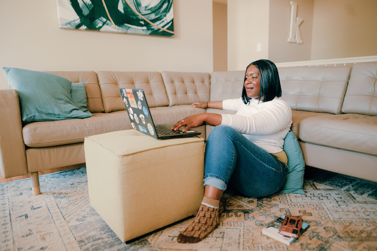 Student sitting on the floor resting against a pillow and couch, while working on a laptop.