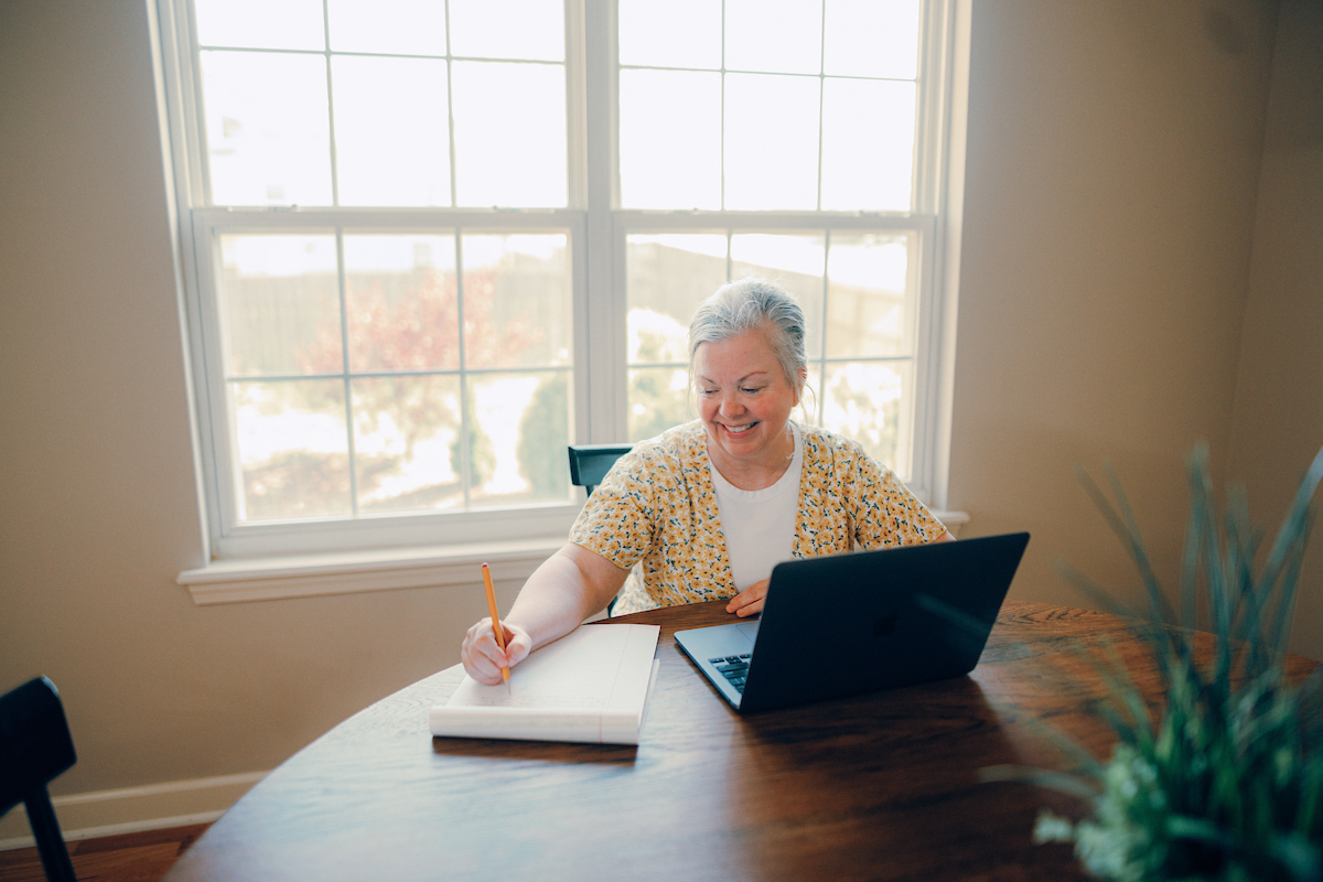 Adult student sitting at dining table in front of window, with a laptop and writing pad in front of her.