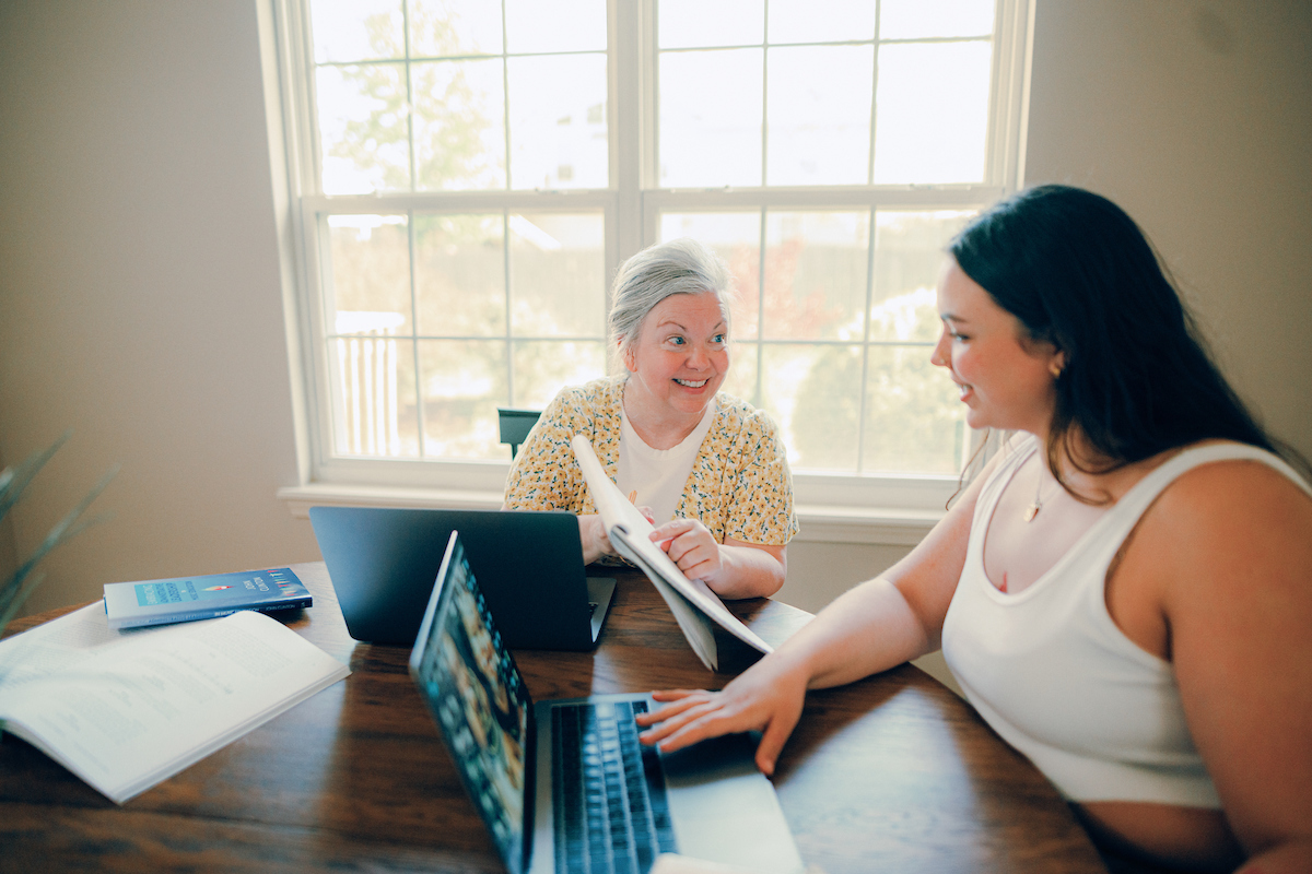 Two online students working together, sitting at a table in front of a window, discussing an item over laptops.