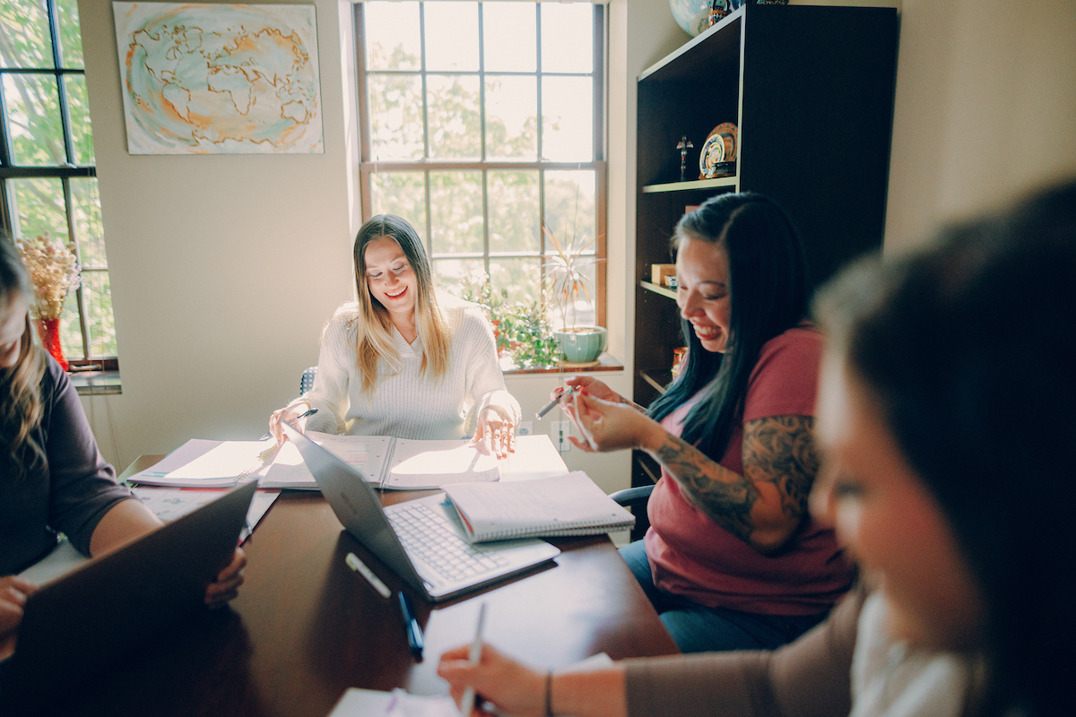 Three students sitting at a table, surrounded by notebooks and laptops as they work together.