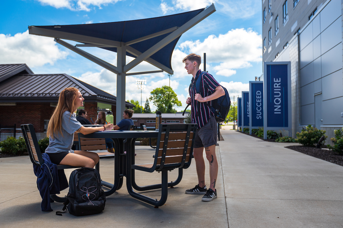 Student seated at a table outside of New Hall talking with another student who is standing and carrying a backpack.