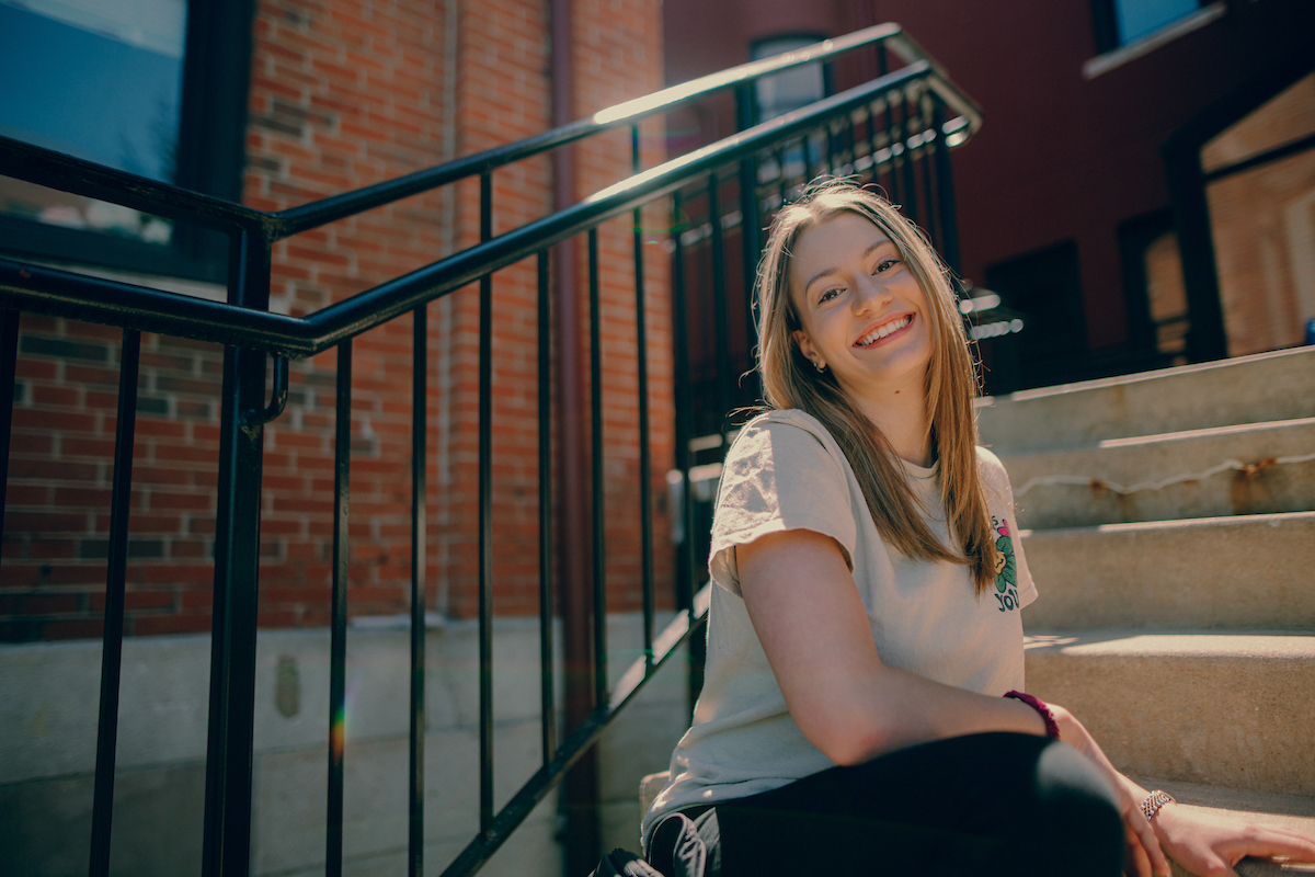 Student sitting on cement steps outside on main campus, turning to smile at the camera.