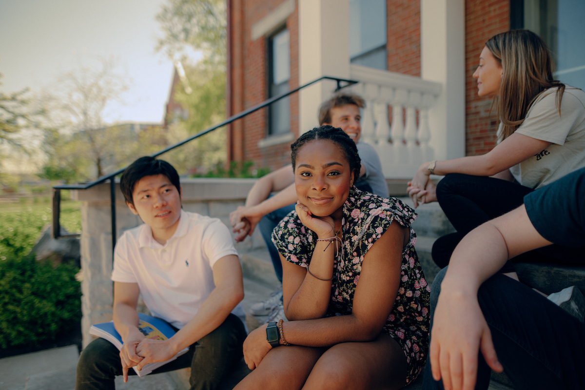 Female student sitting on cement steps, surrounded by other students, with her chin resting on her hand while she smiles directly at the camera.