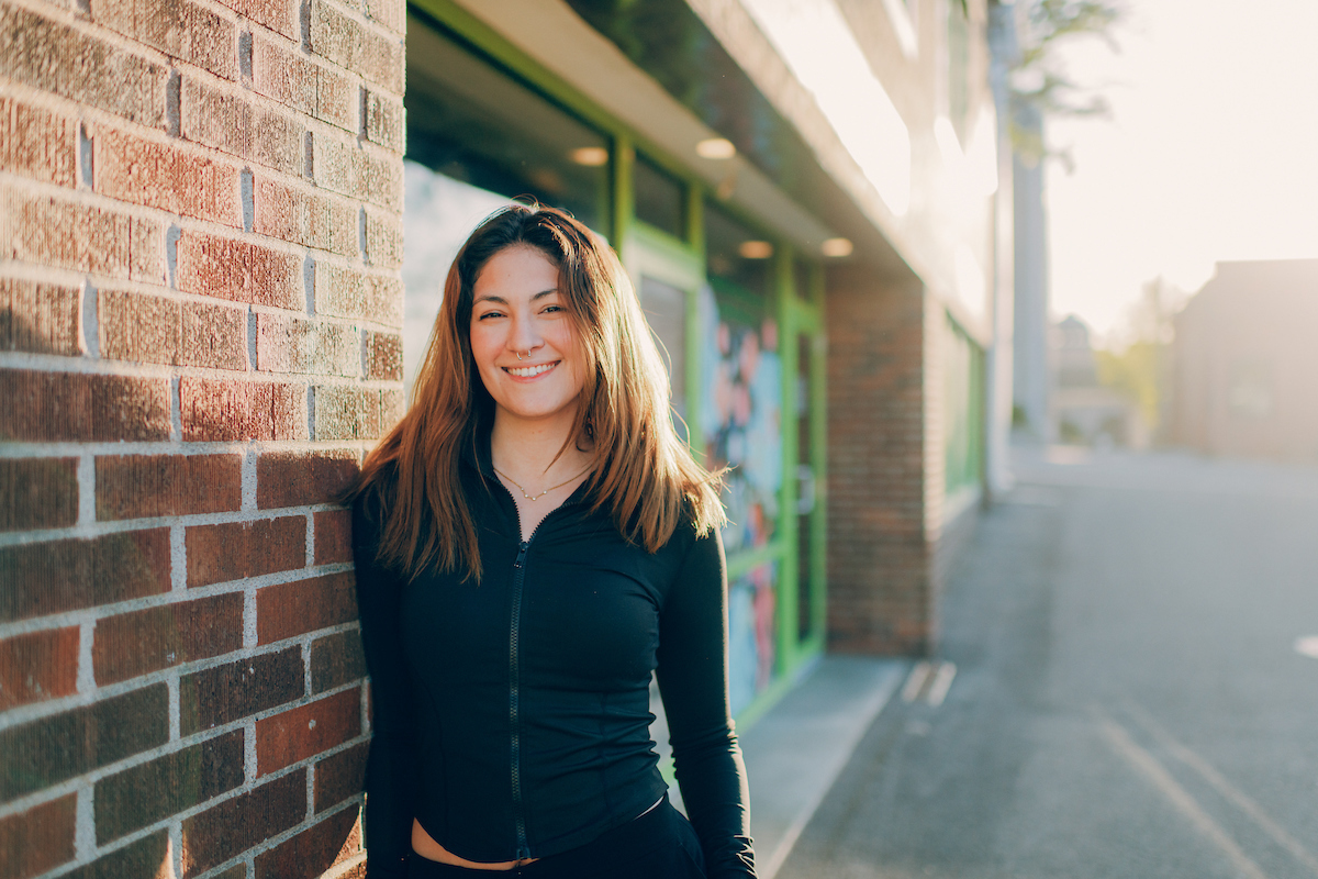 Student with long brown hair, leaning against a brick wall and smiling at the camera.