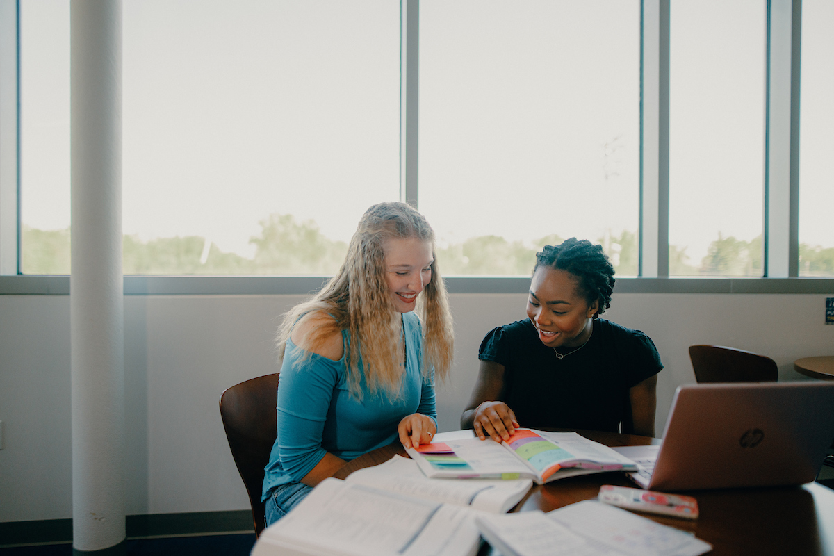 Two students sitting at a table in front of a large window, reviewing the data in a book.