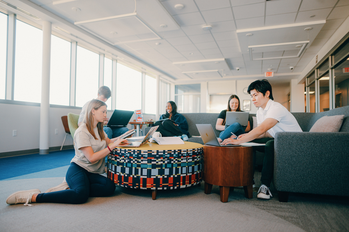 Five students studying together, with four students on a couch with laptops and one student sitting on the floor with her laptop on a table.