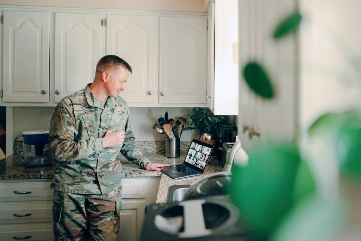 Man in military uniform standing at the kitchen counter reviewing content on his laptop.