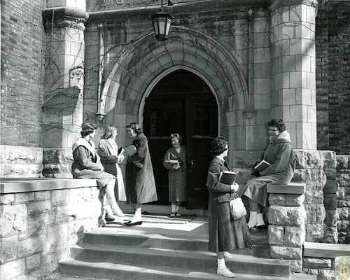 Historical image of several students standing on front cement steps and sitting on cement retaining wall.