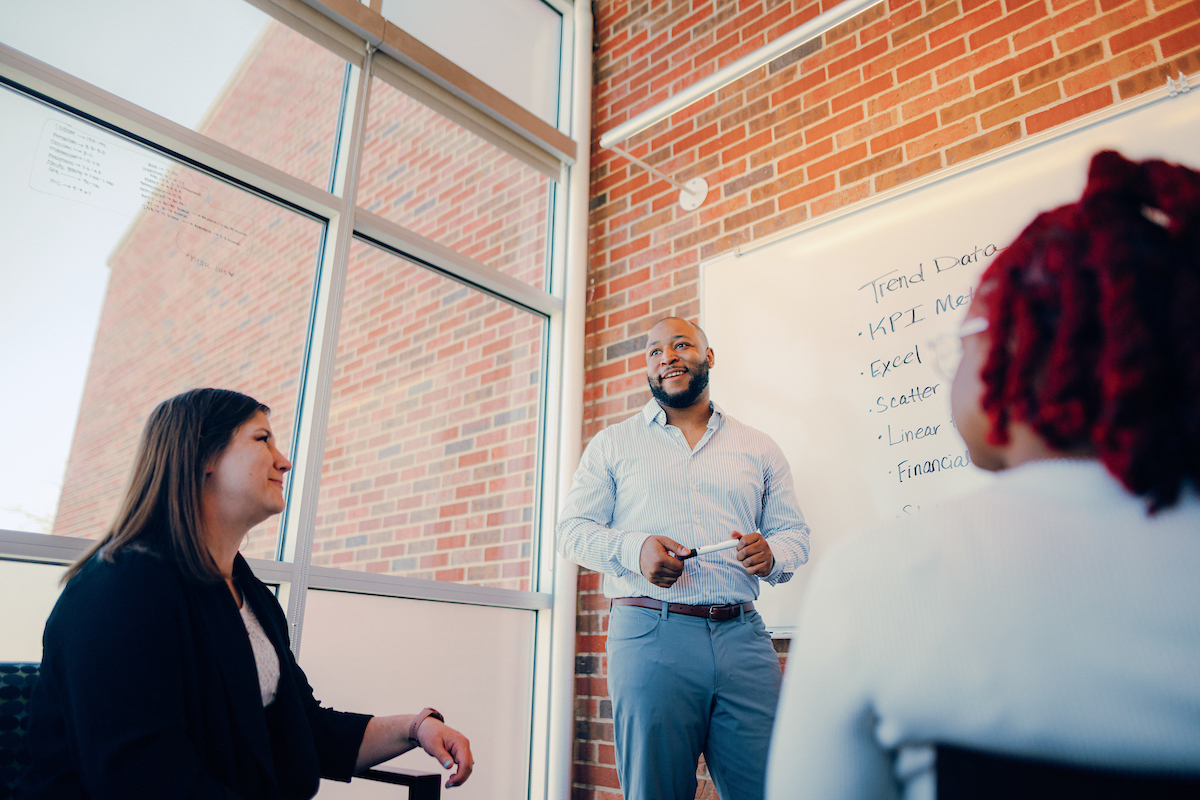 Two people sitting, one person standing in front of a whiteboard, having a discussion.