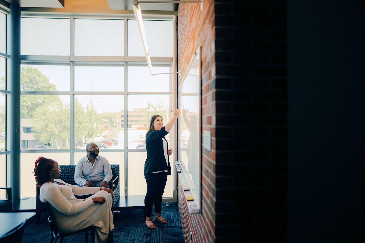 Two people sitting and listening as a person standing points to a whiteboard on the wall.