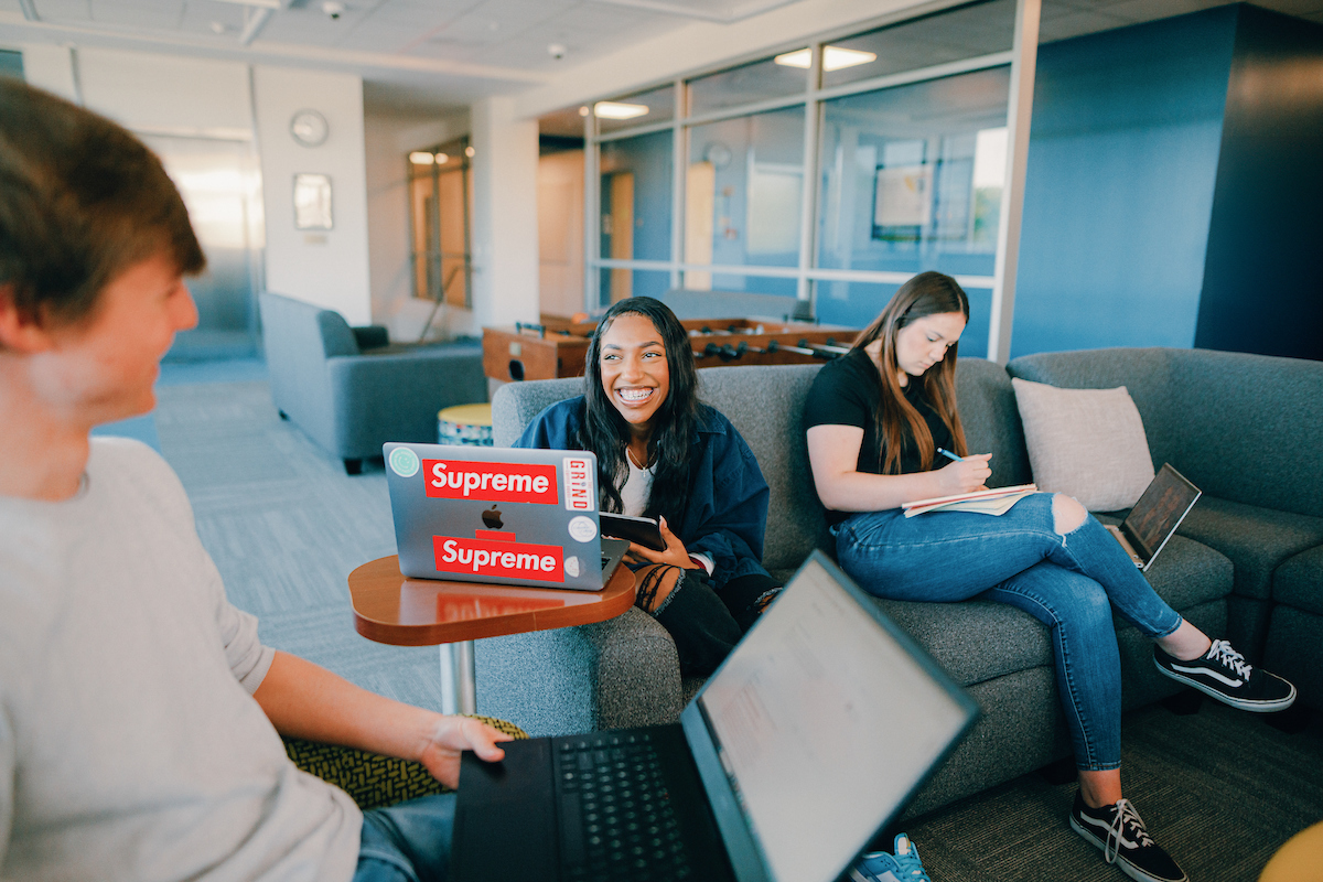 Three students seated in a lounge, working together using laptops and notepads.