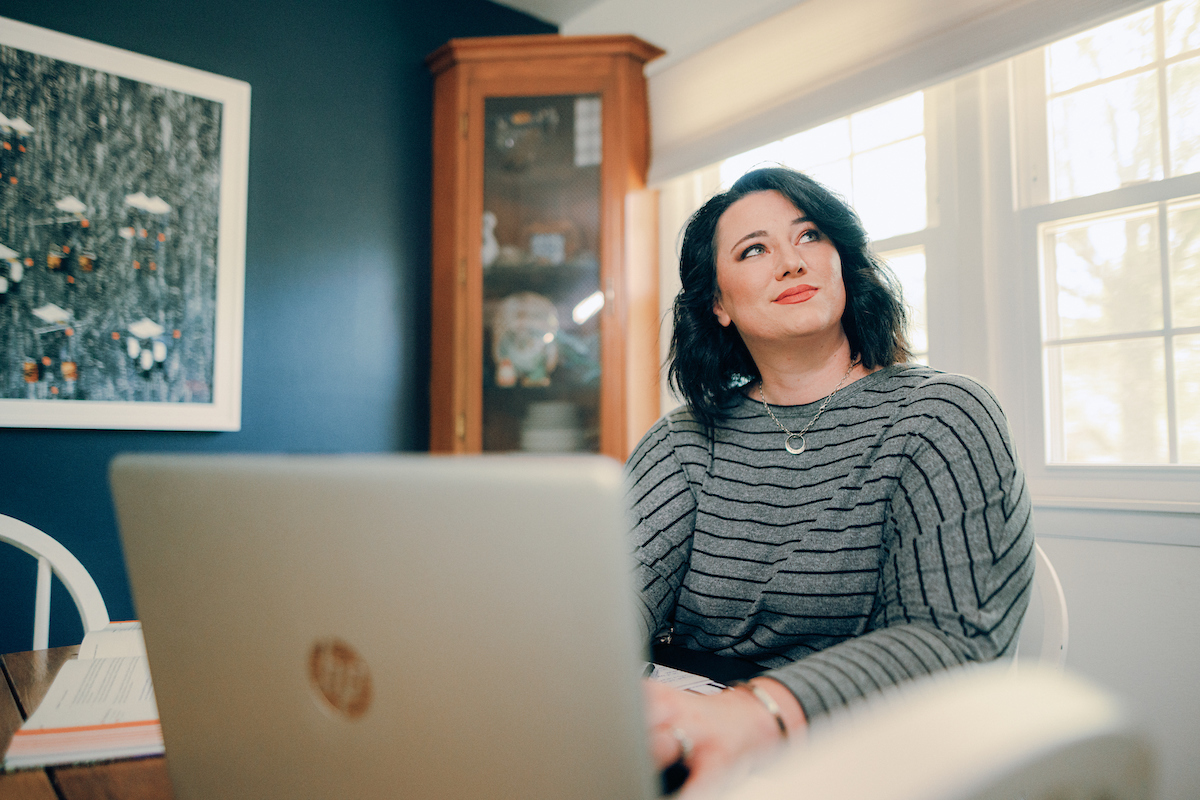 Student sitting at table at home, with laptop in front of her while she gazes up to her left.