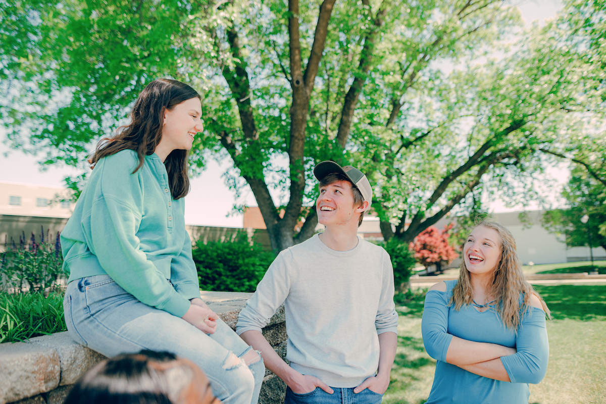 Three students outside in front of a tree, with two students standing and one sitting on a cement retaining wall.